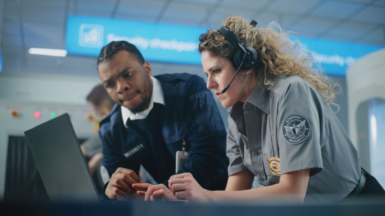 Airport Security Checkpoint: Multiethnic TSA Officers in Headset Using Laptop Computer and Talking. Transportation Security Administration for Travel Safety. In the Background, Airport Staff Working.