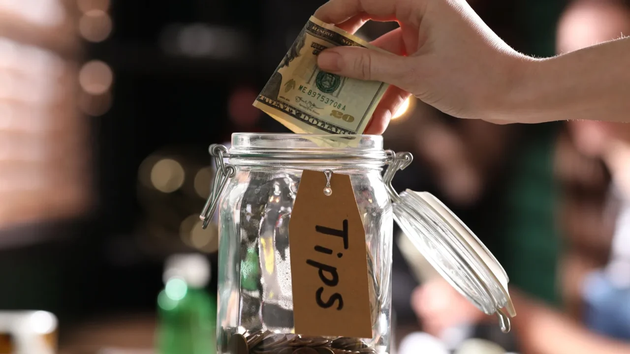 woman putting tips into glass jar on wooden table indoors