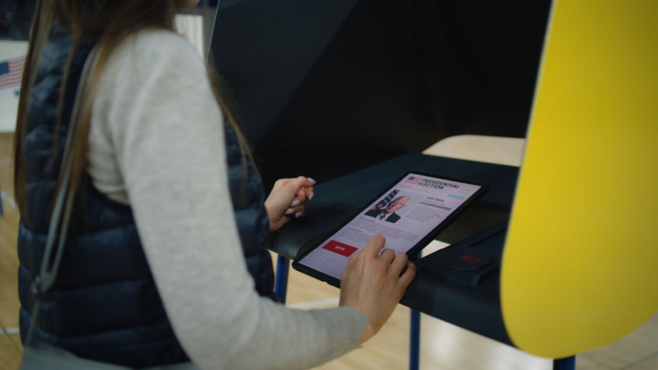 US citizen uses tablet computer for voting. Caucasian woman, female voter thinks and makes choice standing near voting booth at polling station. Presidential Election in the United States of America.