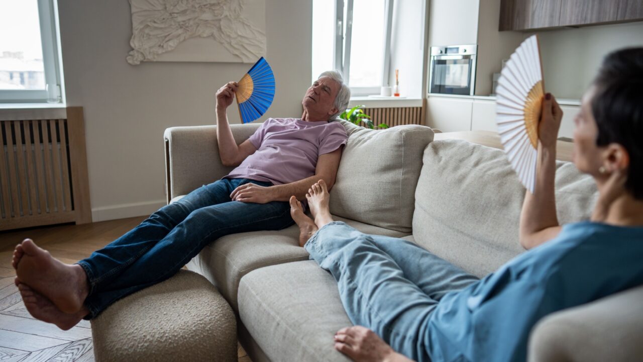 Tired suffering senior couple enduring extreme heat, using fans lounging on couch, too overwhelmed to act. Heatwave impact on elderly, heat discomfort, no air circulation, vulnerable age in summer