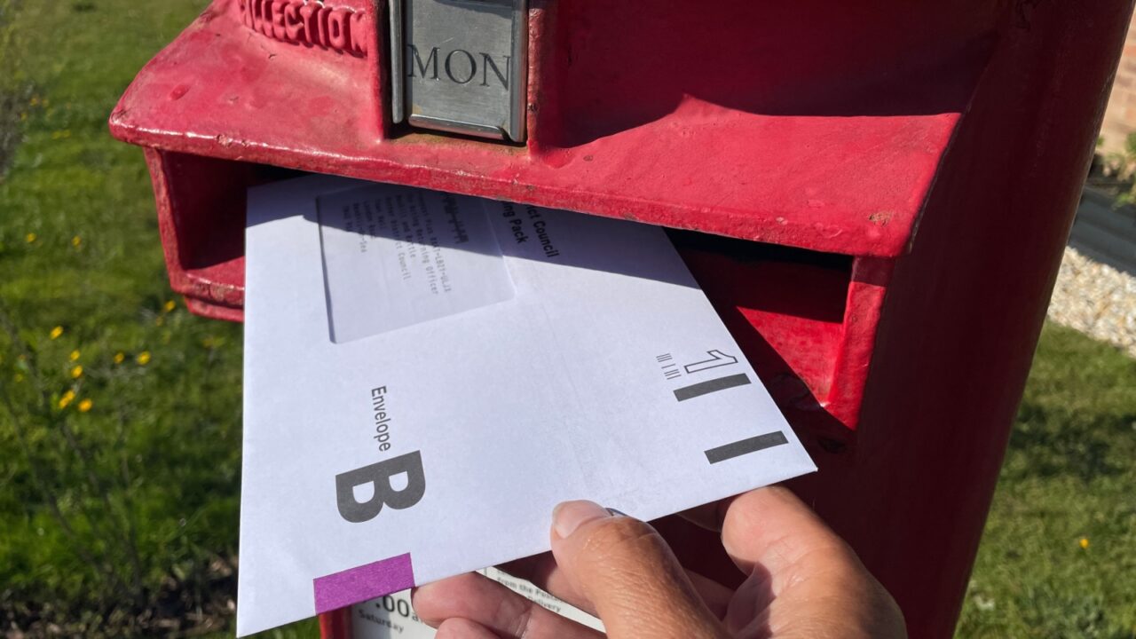 An Envelope B for the United Kingdom General Election postal vote can be seen with a hand putting it in a red post letterbox.First Class