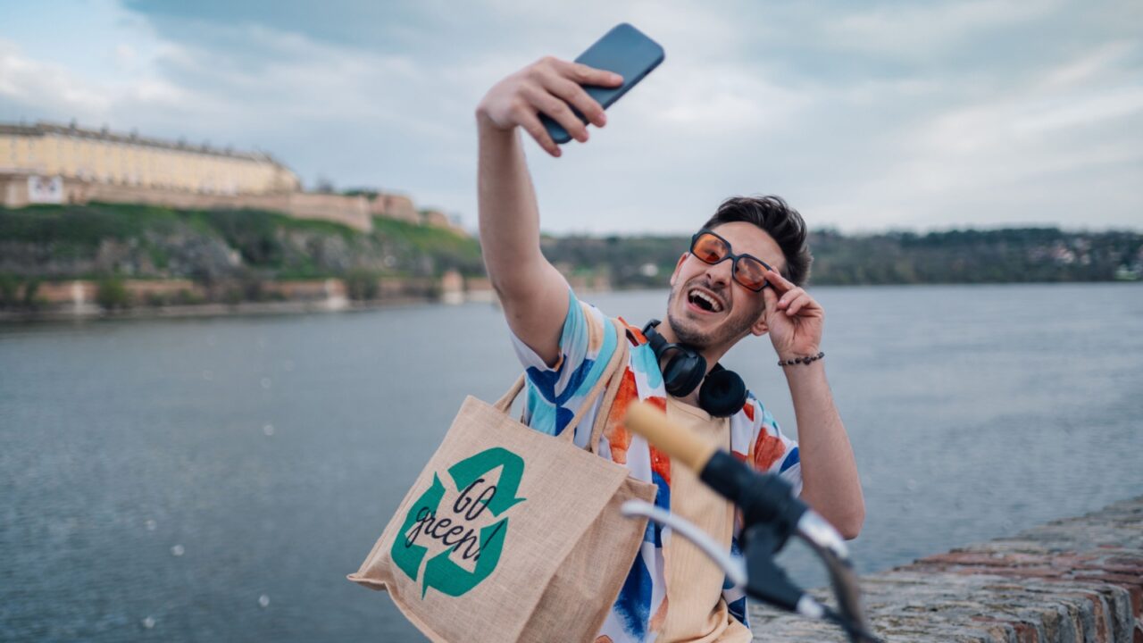 Happy young man with reusable shopping bag and headphones taking selfie near river and promoting sustainable tourism