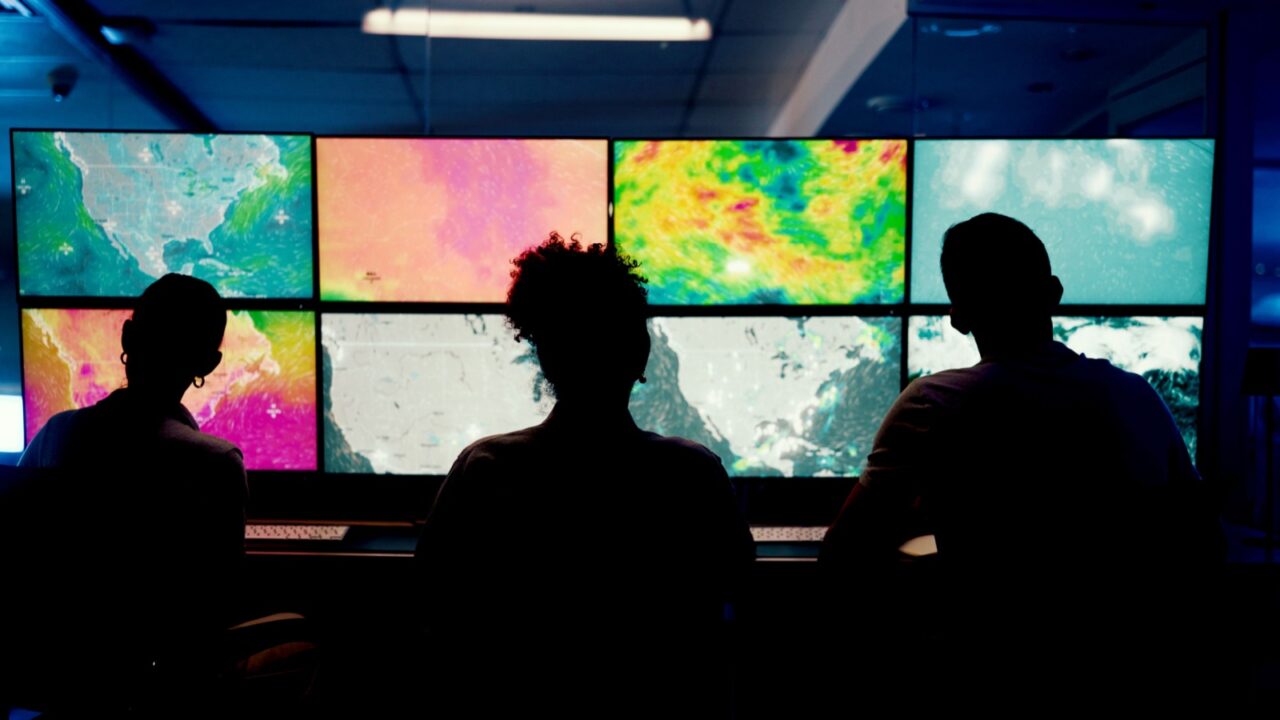 Weather, screen or monitor with people for forecast, satellite maps or teamwork to track storm. Control room, back or scientists with meteorology for broadcast news, report or silhouette at night