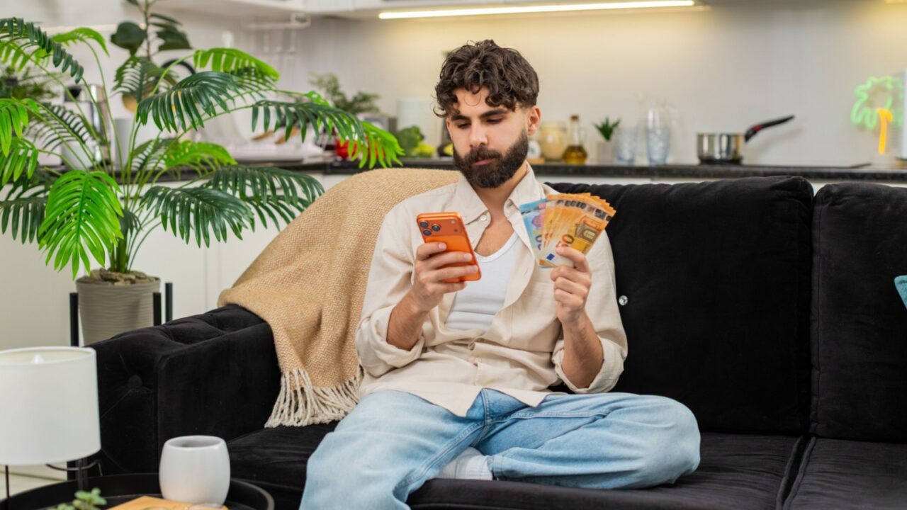 Indian man at home uses smartphone calculator counts euro cash bills plans vacation budget expenses savings. Arabian guy on sofa checks mobile phone compares money with travel cost feels calm focused.